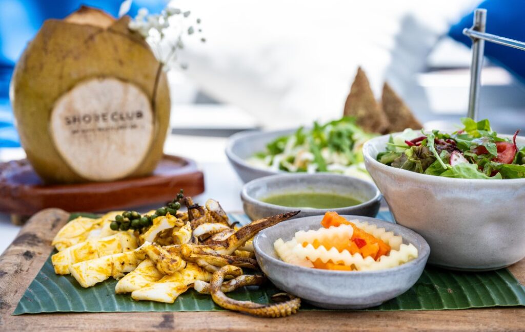 A vibrant spread of Shore Club's food menu, featuring grilled seafood, fresh, healthy salads and gourmet dishes, with an ice cold, fresh coconut on An Bang Beach.